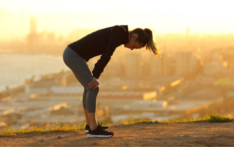 Exhausted female runner at top of hill overlooking city, bending over to catch breath
